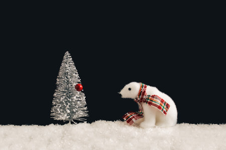 A polar bear with a holiday scarf, white Christmas tree and a red ball in the snow. Black...