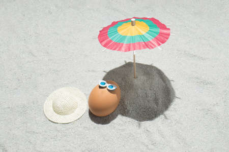 Creative Easter composition with egg, hat and beach umbrella on the sand. Summer and holiday background  idea. Flat lay.の写真素材