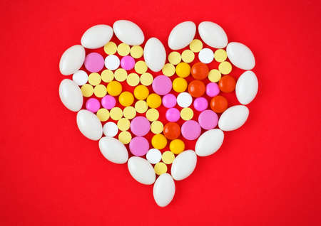 Colorful tablets arranged in a heart shape on red background. Symbol photo of heart disease, medication and pharmaceuticals. Medicine pills or capsules heart shaped on red backgroundの写真素材