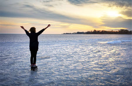 Girl on roller skates standing on a frozen lake and admiring the sunset with hands raisedの写真素材