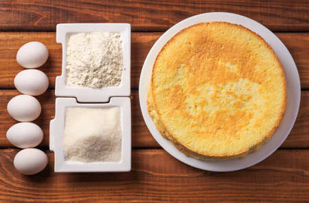 Round homemade golden biscuit cake on a plate, flour, sugar and eggs on wooden table. Ingredients for the cake. The cake and the ingredients from which it was preparedの写真素材