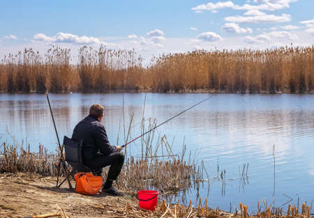 A fisherman with a fishing rod sitting on a chair on the river bank. The concept of a rural getaway. の写真素材