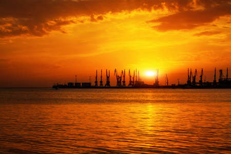 Beautiful landscape with fiery sunset sky and sea. Harbor of Berdyansk during sunrise. Cranes silhouettes against fiery, orange and red sky.の写真素材