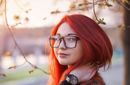 Attractive young girl with red hair, wearing glasses, posing near a tree at sunriseの写真素材