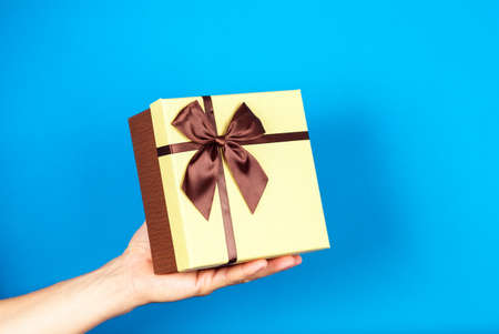 Human hand holding brown ribbon wrapped holiday surprise gift or present box package on blue...