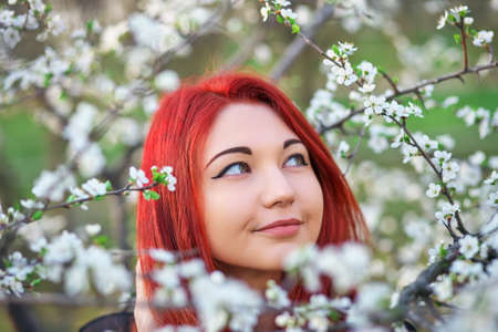 The girl with red hair inhales the fragrance of the flowers of the tree. Outdoor. Spring.