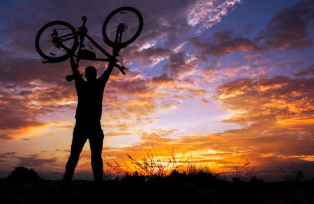 Silhouette the man stand in action lifting bicycle above his head on the meadow with sunset
の写真素材