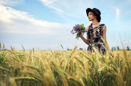 Cute attractive girl with a bouquet of colorful flowers in her hands. Young woman breathes in the scent of plants on wheat field during sunset. Pensive look. Romantic atmosphere.の写真素材