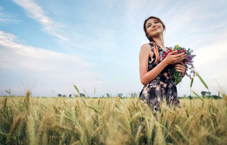 Cute attractive girl with a bouquet of colorful flowers in her hands. Young woman breathes in the scent of plants on wheat field during sunset. Pensive look. Romantic atmosphere.の写真素材