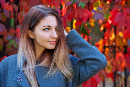 Dreamy beautiful girl with long colorful hair on autumn background of red grape hedge. Inspired woman in gray coat. Autumn euphoria. Female emotional portrait in faded tones.の写真素材