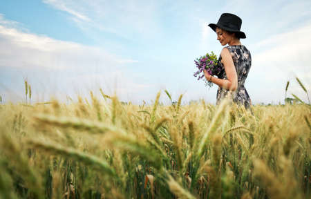 Cute attractive girl with a bouquet of colorful flowers in her hands. Young woman breathes in the scent of plants on wheat field during sunset. Pensive look. Romantic atmosphere.の写真素材