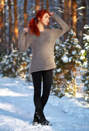 Beautiful charming girl with red hair in the winter park. Pensive, dreamy, loving look. Cold weather. Trees in the snow.の写真素材