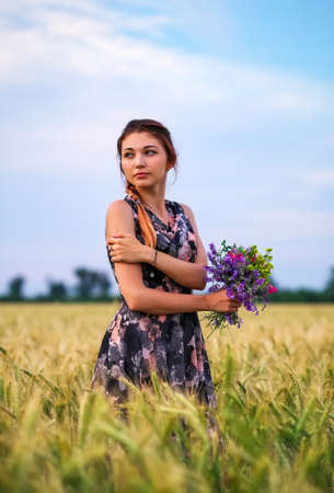 Cute attractive girl with a bouquet of colorful flowers in her hands. Young woman breathes in the scent of plants on wheat field during sunset. Pensive look. Romantic atmosphere.の写真素材