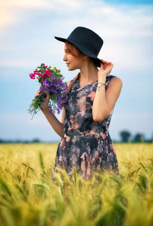 Cute attractive girl with a bouquet of colorful flowers in her hands. Young woman breathes in the scent of plants on wheat field during sunset. Pensive look. Romantic atmosphere.の写真素材
