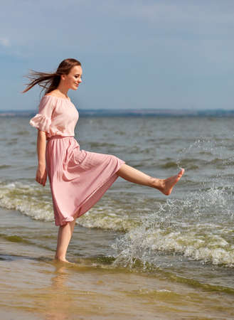 Happy woman in a pink dress walking on the coast of the lake and spraying the water with her feet. A joyful girl stands on the beach of the sea and sprays water with her foot.の写真素材