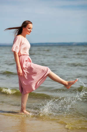 Happy woman in a pink dress walking on the coast of the lake and spraying the water with her feet. A joyful girl stands on the beach of the sea and sprays water with her foot.の写真素材