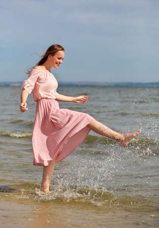 Happy woman in a pink dress walking on the coast of the lake and spraying the water with her feet. A joyful girl stands on the beach of the sea and sprays water with her foot.の写真素材