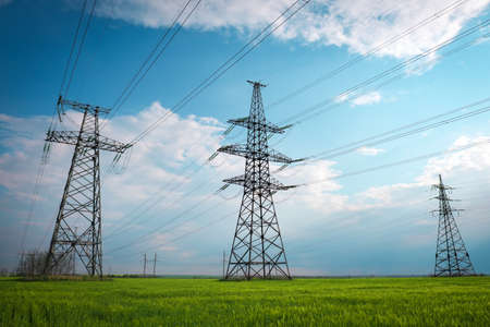 High voltage lines and power pylons in a flat and green agricultural landscape on a sunny day with clouds in the blue sky. Cloudy and rainy. Wheat is growingの写真素材