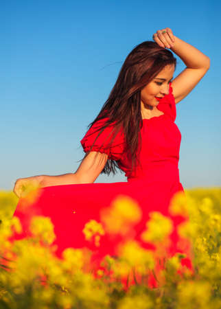 Beautiful young girl in a red dress posing in a field with canola. Happy woman on the nature. Spring season, warm day.の写真素材