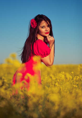 Beautiful young girl in a red dress posing in a field with canola. Happy woman on the nature. Spring season, warm day.の写真素材