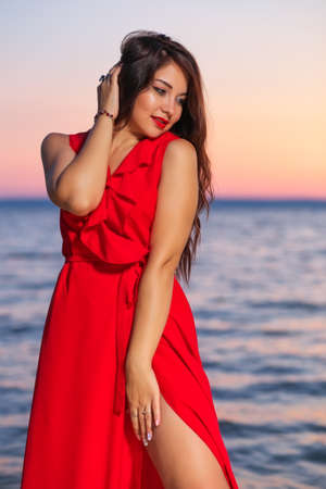 Beautiful brunette girl in a red dress posing on the seashore during sunrise or sunsetの写真素材