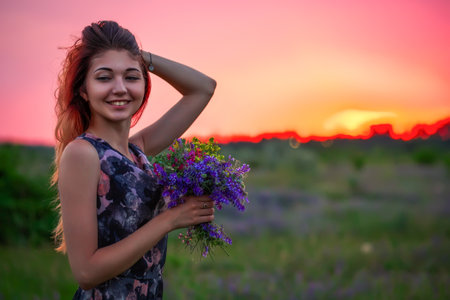 Cute young attractive girl with a bouquet of colorful flowers in her hands. Evening walk in nature during sunset. Pensive look. romantic atmosphere.の写真素材