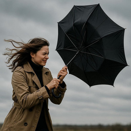 Young woman in trench coat with black umbrella on cloudy sky background.