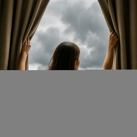 Young woman opening the curtain and looking out the window with cloudy sky