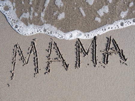 Sand inscription of the word MAMA on a beach, with gentle waves lapping at the shore, creating a serene and heartfelt atmosphere for family moments and memoriesの写真素材