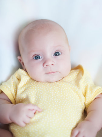 Infant with bright blue eyes wearing a yellow polka dot onesie, lying on a soft surface, showcasing...