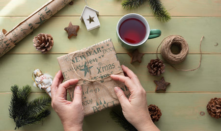 Person's hands are wrapping a gift box with decorative paper, surrounded by pinecones, a cup of tea, and holiday ornaments, evoking a warm festive spiritの写真素材