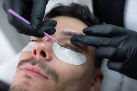 Male client is undergoing eyelash extension application in a modern beauty salon, with a technician...