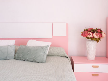 Cozy bedroom featuring a pink bed frame with soft pillows, a textured blanket, and a stylish vase of roses on a matching nightstand, creating a warm and inviting atmosphereの写真素材