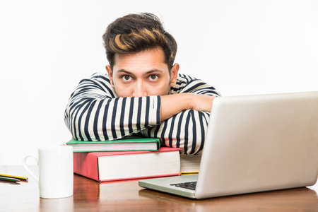 Handsome Indian boy or male college student studying on study table with pile of books, laptop...