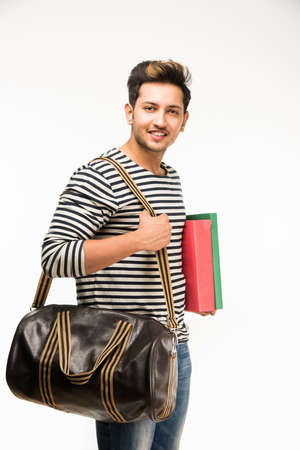 Handsome and young indian Male college student carrying bag on white background while holding college books, laptop or smart phoneの写真素材