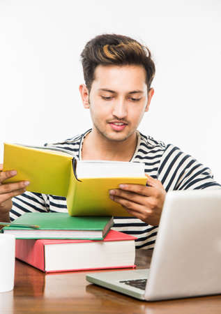 Handsome Indian boy or male college student studying on study table with pile of books, laptop...