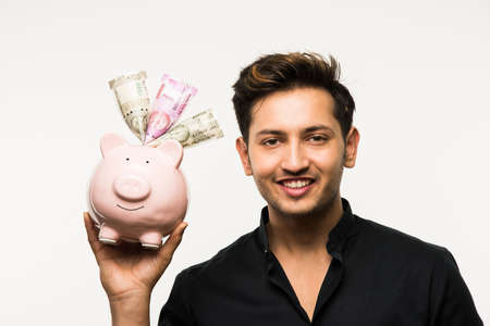 India and finance concept - Handsome Indian man or boy holding pink piggy bank, with different expressions holding indian currency notes of 2000 and 500 or rupee coins, standing over white backgroundの写真素材