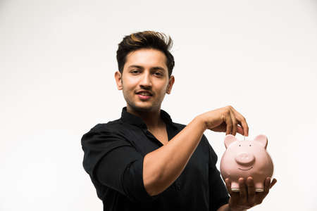 India and finance concept - Handsome Indian man or boy holding pink piggy bank, with different expressions holding indian currency notes of 2000 and 500 or rupee coins, standing over white backgroundの写真素材