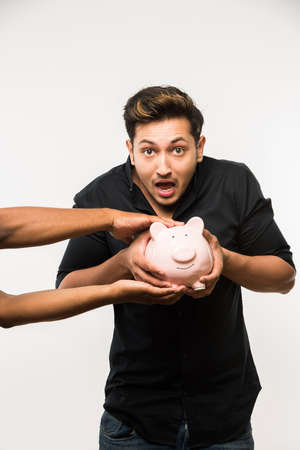 India and finance concept - Handsome Indian man or boy holding pink piggy bank tightly and protecting his saving from theft, standing over white backgroundの写真素材