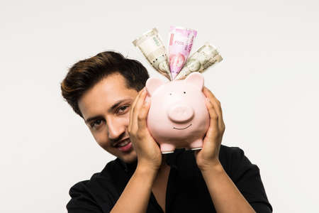 India and finance concept - Handsome Indian man or boy holding pink piggy bank, with different expressions holding indian currency notes of 2000 and 500 or rupee coins, standing over white backgroundの写真素材