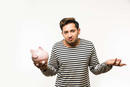 India and finance concept - Handsome Indian man or boy holding pink piggy bank, with different expressions holding indian currency notes of 2000 and 500 or rupee coins, standing over white backgroundの写真素材