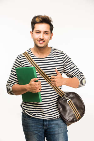 Handsome and young indian Male college student carrying bag on white background while holding college books, laptop or smart phoneの写真素材