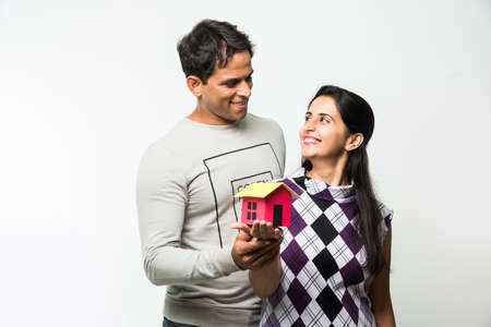 stock photo of Indian smart and cheerful / happy couple holding 3D paper house model and keys, standing isolated over white background, asian couple and real estate conceptの写真素材