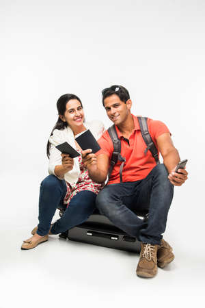 indian couple sitting over packed suitcase, waiting for departure with passports in hand or taking selfie. isolated over white backgroundの写真素材