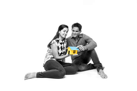 stock photo showing side angle of Indian or asian smart and cheerful / happy couple holding 3D paper house model and sitting isolated over white background, asian couple and real estate concept.の写真素材