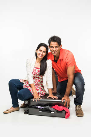 stock photo of indian young Couple packing for holiday, trying to close suitcase full with clothes, sitting isolated over white backgroundの写真素材