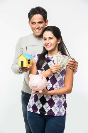 Indian smart and cheerful / happy couple holding currency notes, Pink Piggy bank and 3D paper house model isolated over white background, asian couple and real estateの写真素材
