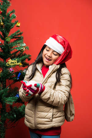 Indian little cute girl holding or blowing snow on christmas with christmas tree and red backgroundの写真素材