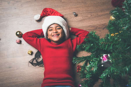Cute Indian kid girl playing under christmas tree, lying on wooden floor in room. Wearing trendy red knitted sweater, jeans with santa hat and white snow. Looking at camera. Top view.の写真素材