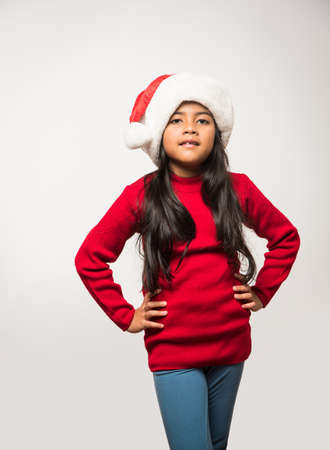 cheerful Indian girl child with santa hat posing or holding golden decorative balls over white or red or blue background on christmasの写真素材